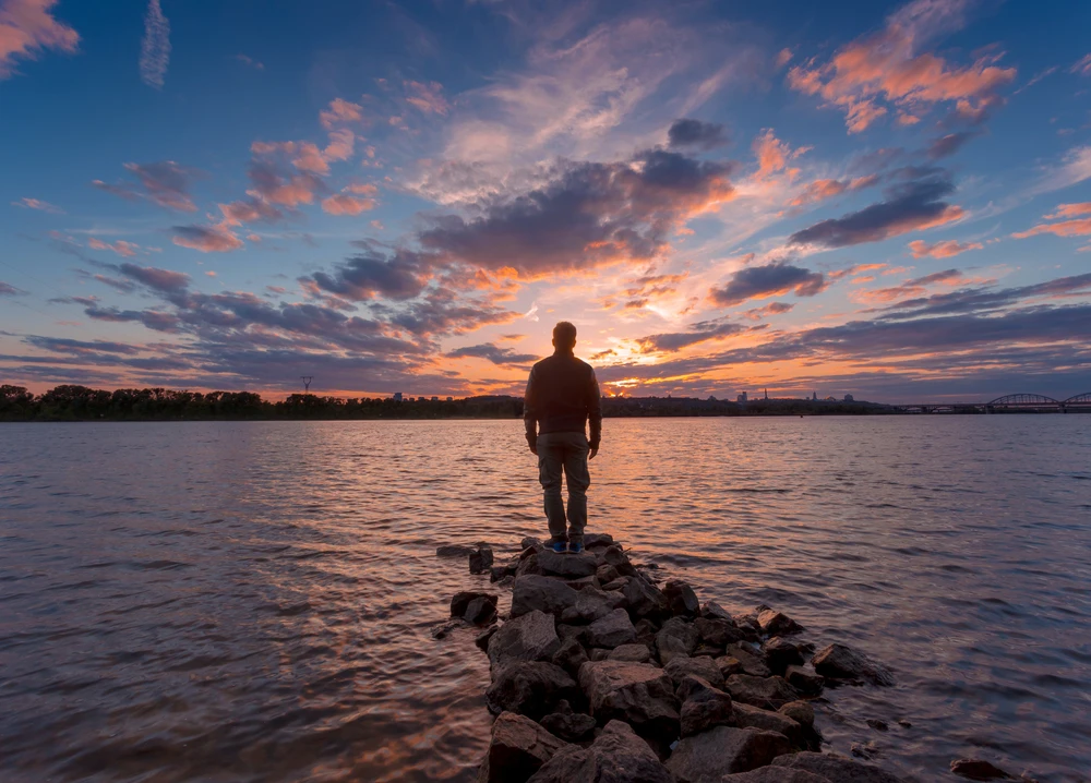 Man staat op rotsen aan het water naar zonsopkomst of zonsondergang te kijken.