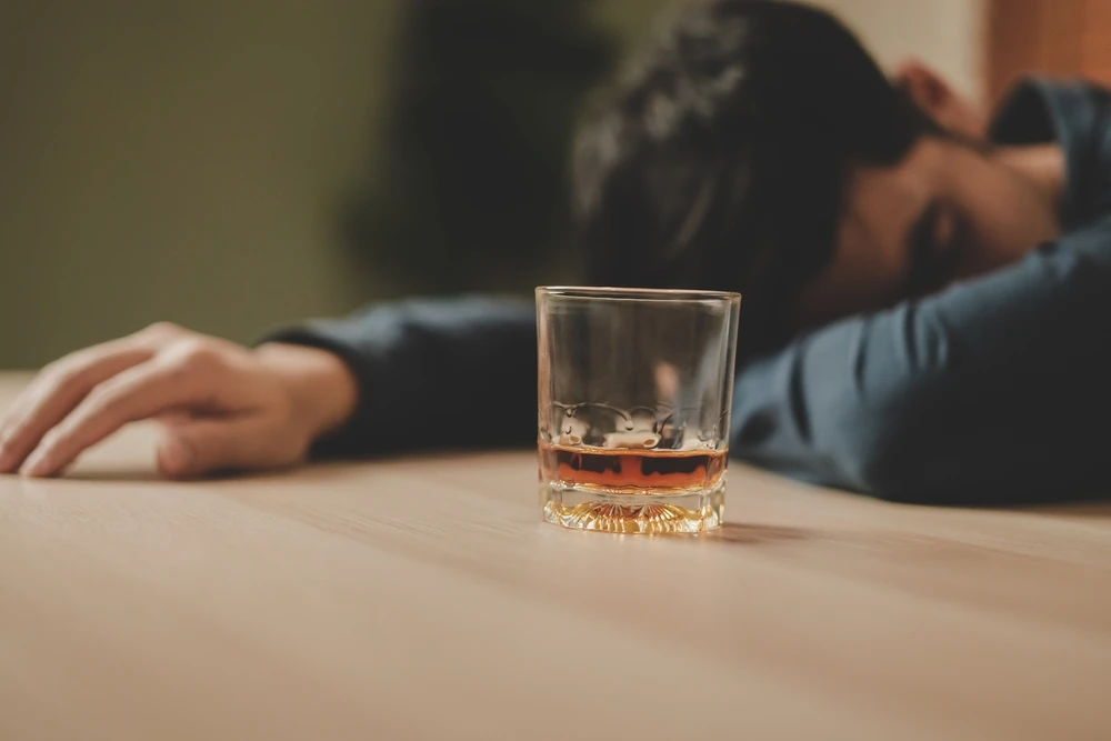Man ligt met zijn hoofd op tafel te slapen naast een bijna leeg glas alcohol.