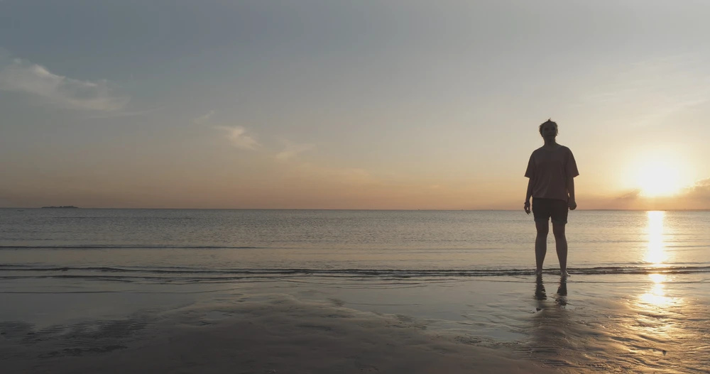 Persoon met korte broek en t-shirt staat wat verloren tegen de zon in gefotografeerd met de voeten in de zee.