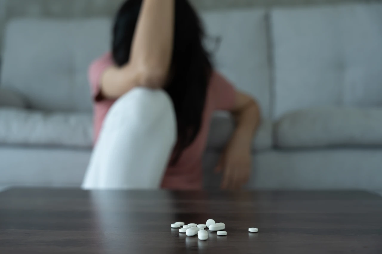 Vrouw met gezicht verborgen achter haren zit wanhopig op de grond met pillen op tafel. 