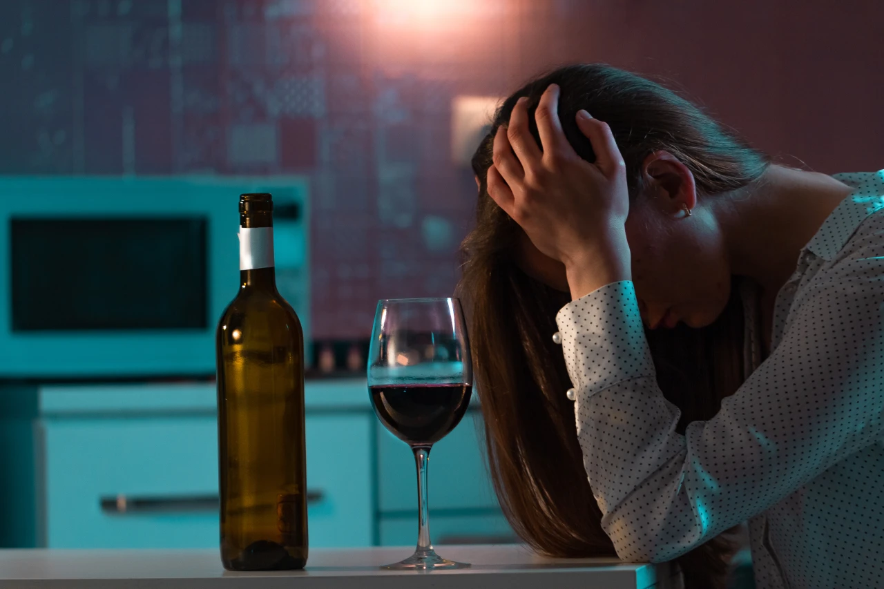 vrouw-met-zorgen-en-wijn Vrouw aan eettafel maakt zich zorgen, heeft het hoofd in handen; wijnfles en glas op tafel.