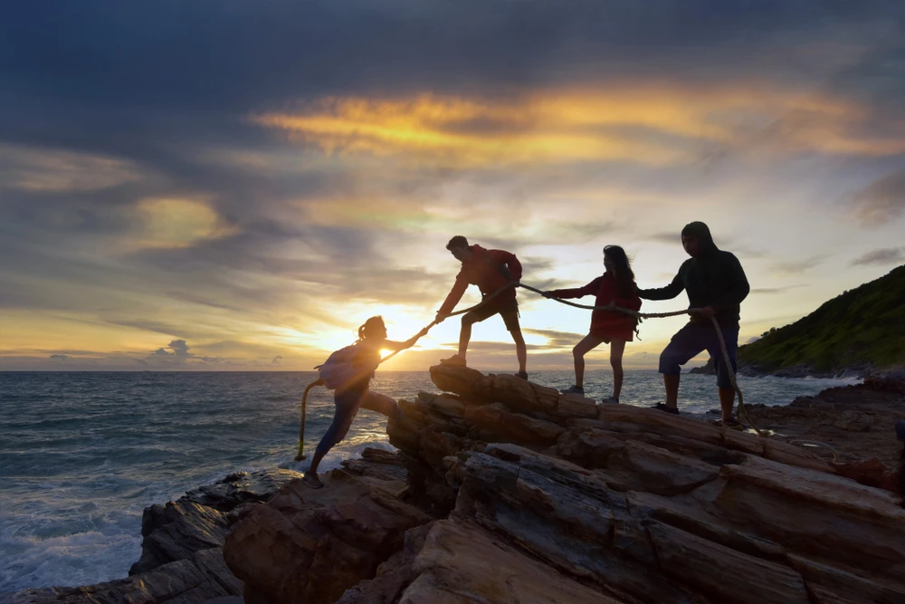 Vier mensen helpen elkaar om met een touw tegen rotsige zeekust op te klimmen. 